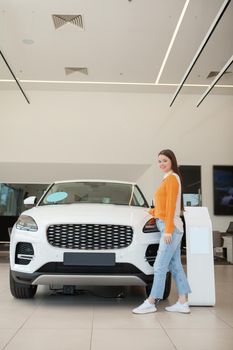 Vertical full length shot of a beautiful young woman standing near her newly bought car at auto dealership