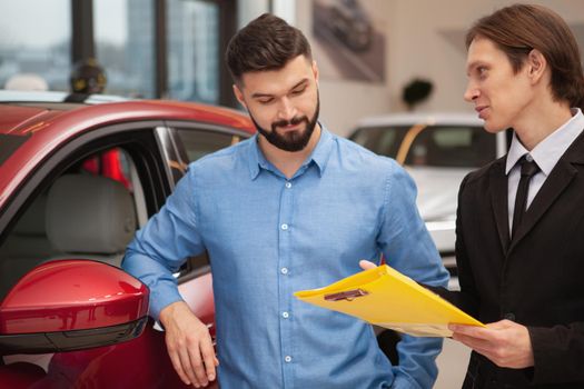 Young male car dealer talking to his male customer. Attractive bearded man buying new automobile, talking to the manager at cars dealership