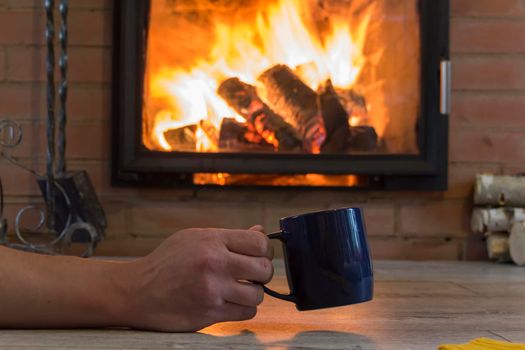 A man's hand holds a mug with a hot drink near the fireplace with a fire. Keep warm in a warm house in winter..