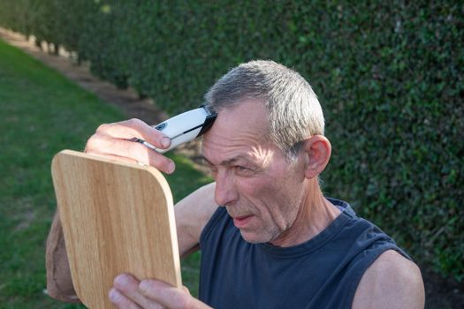 a gray-haired middle-aged man shaves his hair with a clipper in a garden on a green lawn and looks into a table mirror. High quality photo