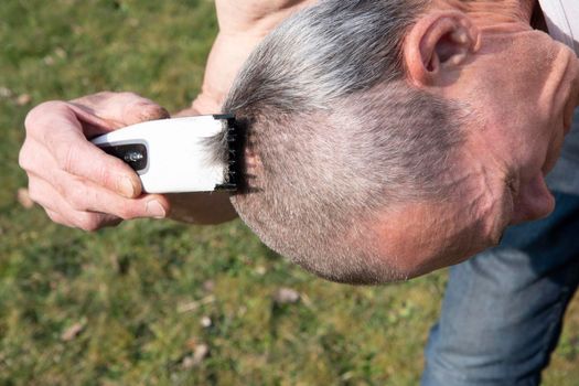 a gray-haired middle-aged man shaves his hair with a clipper in a garden on a green lawn, High quality photo