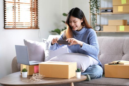 Asian woman photographing parcel boxes with smartphone to deliver goods to customers online on the Internet, starting a small business owner's e-commerce concept