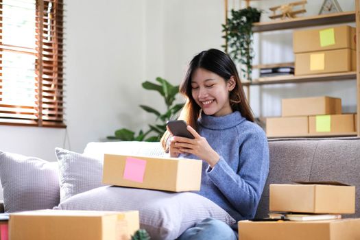 Happy young Asian woman entrepreneur, Smile for sales success after checking order from online shopping store in a smartphone at home office, Concept of merchant business online and eCommerce.