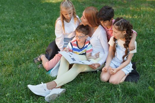 Female teacher and her young students sitting on the grass, reading a book