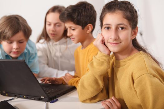 Cute Arab schoolgirl smiling to the camera, her friends using laptop at school on background