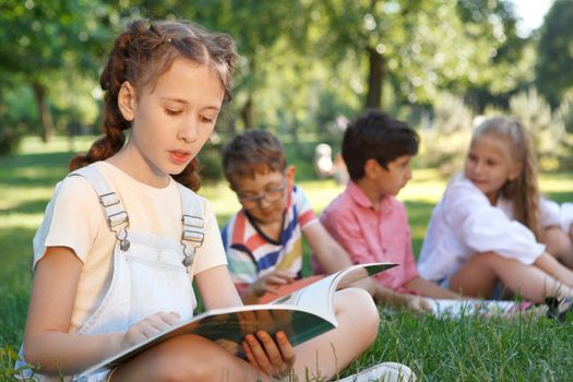 Young girl reading a book at the park, her friends relaxing on the grass on background