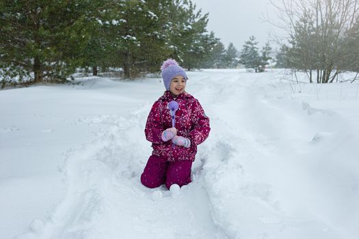 A little girl , sits in the snow, makes snowballs using a plastic sculpting tool. Close up