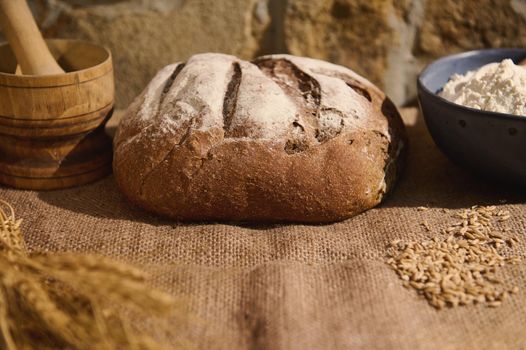 Food composition with a loaf of homemade fresh baked wheaten sourdough bread, wheat spikelets and baking ingredients on a burlap tablecloth, against a stone wall background with copy advertising space