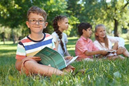 Charming little boy smiling to the camera, while reading a book, sitting on the grass