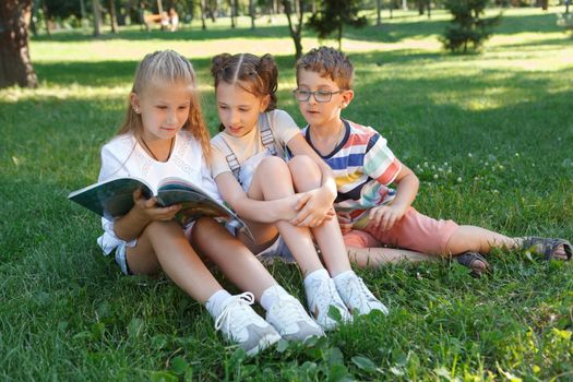 Three children reading a book together on the grass at the park