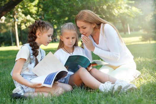 Beautiful female teacher whispering something to her little student while reading a book in the park