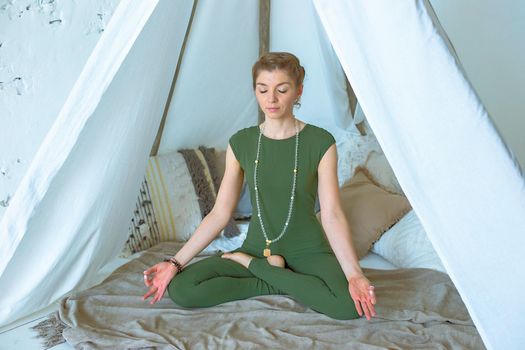 Beautiful and slender girl in green sportswear, meditates or practices yoga at home under a canopy of fabric