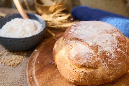 Selective focus on a loaf of homemade whole grain sourdough bread, freshly baked according to family tradition recipe, on a wooden board, bowl with flour and spikelets of wheat. Artisanal bakery store