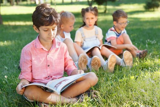 Young boy reading a book, sitting on the grass at the park, his friends talking on background