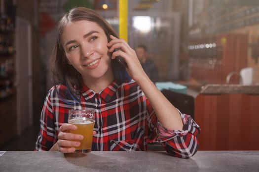 Young happy woman talking on the phone while drinking craft beer at the pub
