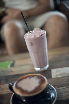 Fruit milkshake standing alongside a cup of cappuccino on a restaurant table, legs of a person seated at the table visible in the background