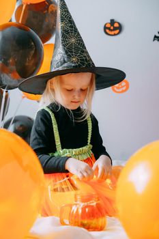 Children's Halloween - a girl in a witch hat and a carnival costume with airy orange and black balloons at home. Ready to celebrate Halloween.