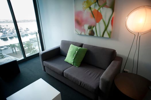 Interior decor of a compact city apartment with a modern lounge with an illuminated standard lamp alongside a grey upholstered couch with modern artwork and a view window
