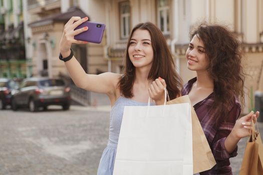 Happy female friends taking selfies with shopping bags, while wandering on city streets, copy space. CHeerful female shoppers using smart phone, taking photos after shopping