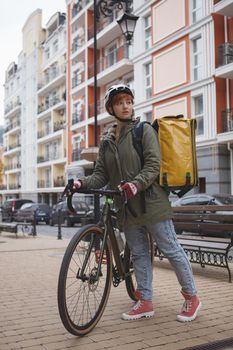 Vertical full length shot of a female courier wearing cycling helmet and delivery backpack walking with her bicycle