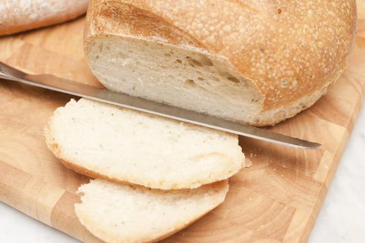 Round loaf of fresh white bread with a knife and two cut slices on a wooden bread board