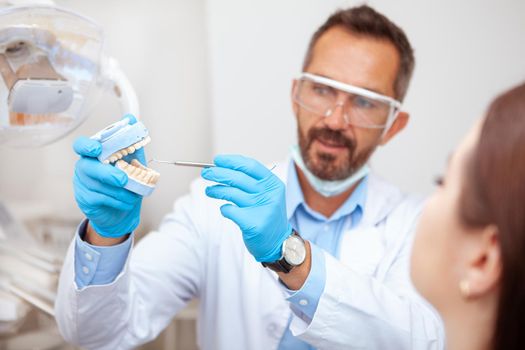 Experienced mature male dentist explaining dental hygiene to his female patient, holding teeth mold