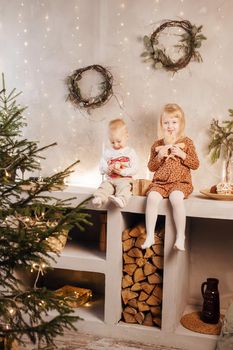 Little brother and sister play on Christmas eve in a beautiful house decorated for the New Year holidays. Children are playing with a Christmas gift. Scandinavian-style interior with live fir trees and a wooden staircase.