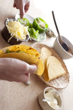 Man filling a taco with fresh ingredients reaching for grated cheese and sliced green pepper on plates on the table