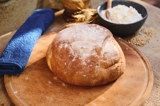 Still life with a loaf of homemade whole grain sourdough bread, freshly baked according to family traditions, on a wooden board, a blue terry towel and a bowl with wheat flour. Artisanal bakery store