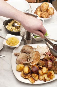 Man serving a family roast dinner using a pair of tongs to dish up carved chicken or turkey from a table laid with vegetables and sauces