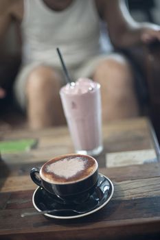 Cup of cappuccino and a milkshake on a table in a restaurant or cafeteria with a persons legs visible behind