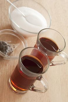 Two cups of hot sweet black tea in glass mugs viewed high angle on a wooden table with the sugar bowl behind and used teabags in a dish