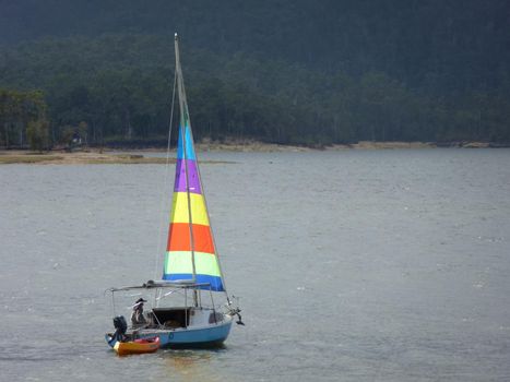 Small sailboat with a colorful sail sailing offshore with a lone sailor towing a dinghy for access to the shore from a mooring