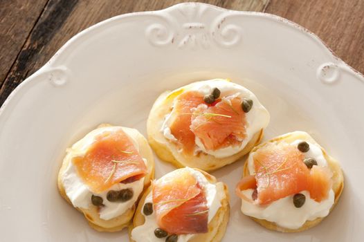 Capers and raw fish blini appetizers on white porcelain decorative plate over wooden table