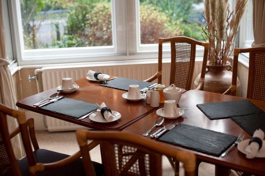 Dining table laid for breakfast with three place settings including coffee mugs beneath a window overlooking the garden