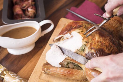 Man carving the Christmas turkey with a carving knife and fork at the table with a gravy sauce boat and roast potatoes alongside