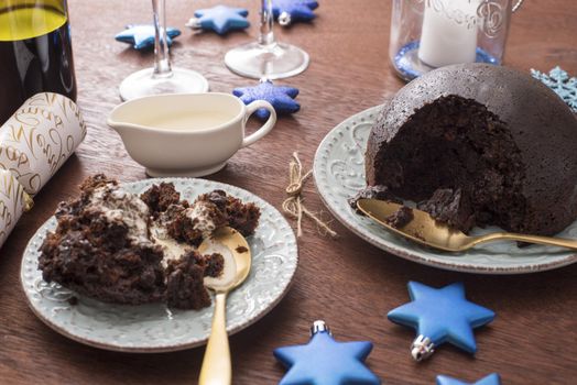 Serving of homemade fruity Christmas pudding topped with brandy cream on a festive dinner table with blue themed decorations