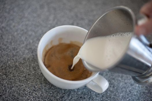 Hand pouring frothy steamed milk from a metal jug into a cup of freshly brewed espresso coffee, high angle view