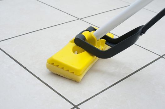 Mopping the white tiled kitchen or bathroom floor using a colorful yellow plastic squeegee in a household chores, cleanliness and hygiene concept