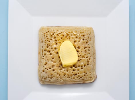 Overhead view of white square bowl with a golden crumpet and a dab of butter on top