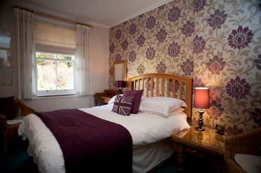 Cozy bedroom with a comfortable wooden slatted bed with a lamp burning on the bedside table and bold wallpaper with a big motif