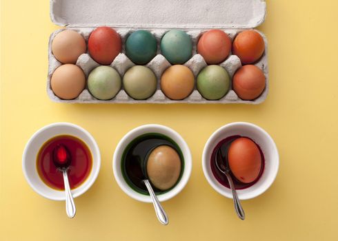 Preparing dyed colored eggs for Easter steeping them in three bowls of dye with a full cardboard carton of finished eggs alongside, overhead view