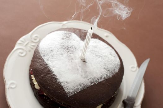 A chocolate birthday cake for a loved one with a white icing sugar heart shape on top and a single extinguished candle