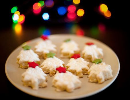 Plate of decorative Christmas biscuits in the shape of Christmas trees with stars standing in front of colourful festive party lights on a black background