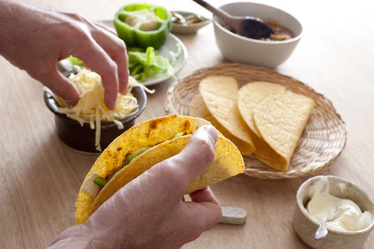 Man preparing Mexican tacos for lunch filling a hard shell with grated cheese, savory mince meat , sour cream and green pepper