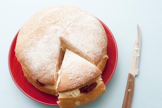 Delicious fresh sponge cream cake with strawberry jam sliced for tea on a red plate, high angle view with knife alongside