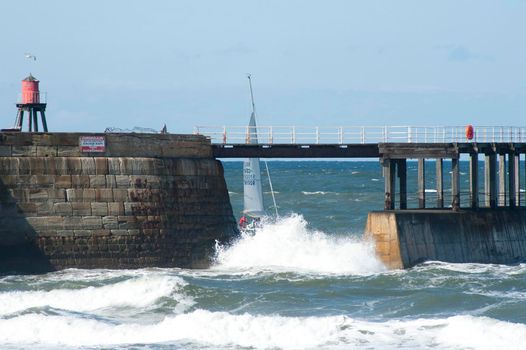 Sailing yacht passing through the sheltered waters behind the Whitby breakwater and stone pier with turbulent surf in the foreground