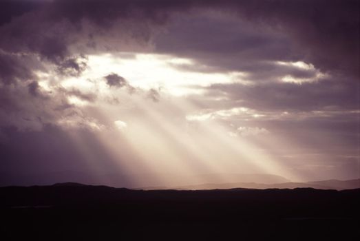 Sunbeams shining through a hole in the dense cloud cover in a stormy grey sky throwing diagonal rays onto the silhouetted hills below