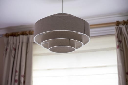 A white, contemporary ceiling light shade hanging from the interior of a modern home with hanging curtains.