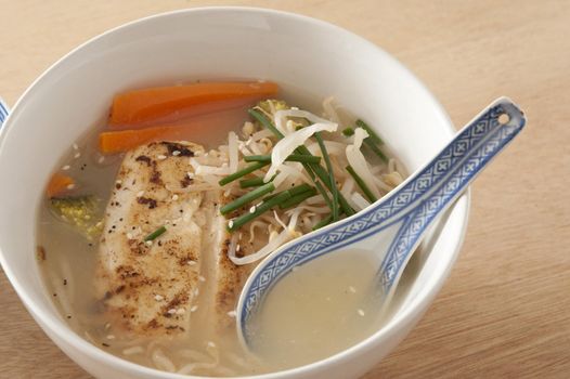 Close up on small white bowl of warm baked fish and noodle soup with garlic, chives, carrots and other herbs on wooden table